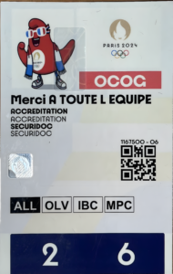 Carte d'accréditation officielle Paris 2024. La mascotte Phryge olympique rouge, portant des lunettes de soleil tricolores, une écharpe bleu-blanc-rouge et un t-shirt avec le logo des JO, est représentée en haut à gauche. Logo Paris 2024 avec flamme et anneaux olympiques. Mention « Merci à toute l'équipe » en majuscules, textes répétés « ACCREDITATION » et « SECURIDOC ». Code QR, numéro 1167500-06, zones d'accès ALL - OLV - IBC - MPC, et indication 2 / 6 en bas